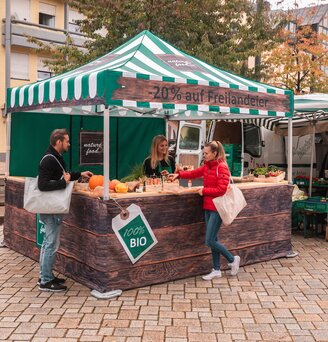 Der Marktstand ist vollflächig bedruckt und kommt auf dem Wochenmarkt und Bauernmarkt zum Einsatz. 2 Kunden sehen sich die Produkte an.