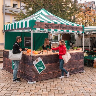 Il gazebo da mercato è completamente stampato e viene utilizzato al mercato settimanale e al mercato degli agricoltori. 2 clienti guardano i prodotti.