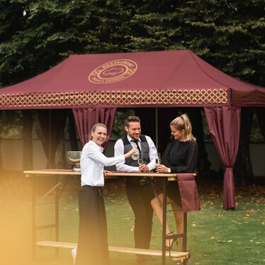 gazebo pieghevole elegante 8x4 m bordeaux con decorazioni oro e tende decorative copripalo. Il gazebo si trova in un prato e serve per il ricevimento di un evento elegante mentre delle persone bevono dello spumante servite da una cameriera