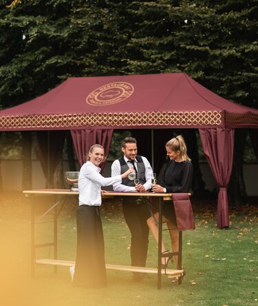 gazebo pieghevole elegante 8x4 m bordeaux con decorazioni oro e tende decorative copripalo. Il gazebo si trova in un prato e serve per il ricevimento di un evento elegante mentre delle persone bevono dello spumante servite da una cameriera