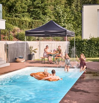 Der schwarze Faltpavillon steht am Pool im Garten. Darunter sitzen Freunde im Schatten und feiern eine Pool- und Gartenparty.