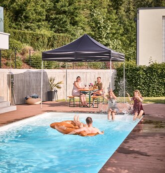 Der schwarze Faltpavillon steht am Pool im Garten. Darunter sitzen Freunde im Schatten und feiern eine Pool- und Gartenparty.