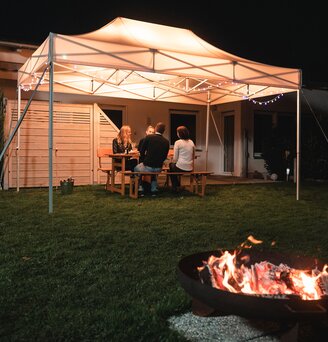 Il gazebo da giardino di 4,5x3 m si trova in giardino al buio. I 4 ospiti siedono sotto di essa. Davanti al gazebo c'è un camino.