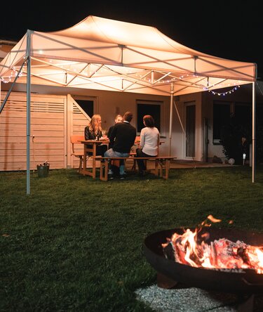 Il gazebo da giardino di 4,5x3 m si trova in giardino al buio. I 4 ospiti siedono sotto di essa. Davanti al gazebo c'è un camino.