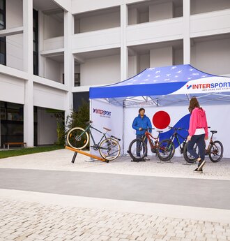 Il gazebo promozionale stampato di "Intersport" si trova nell'area esterna della fiera. Sotto il gazebo pieghevole di 4,5x3 m si trovano il venditore e le sue biciclette.