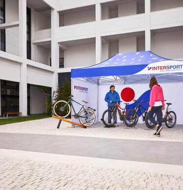 Il gazebo promozionale stampato di "Intersport" si trova nell'area esterna della fiera. Sotto il gazebo pieghevole di 4,5x3 m si trovano il venditore e le sue biciclette.