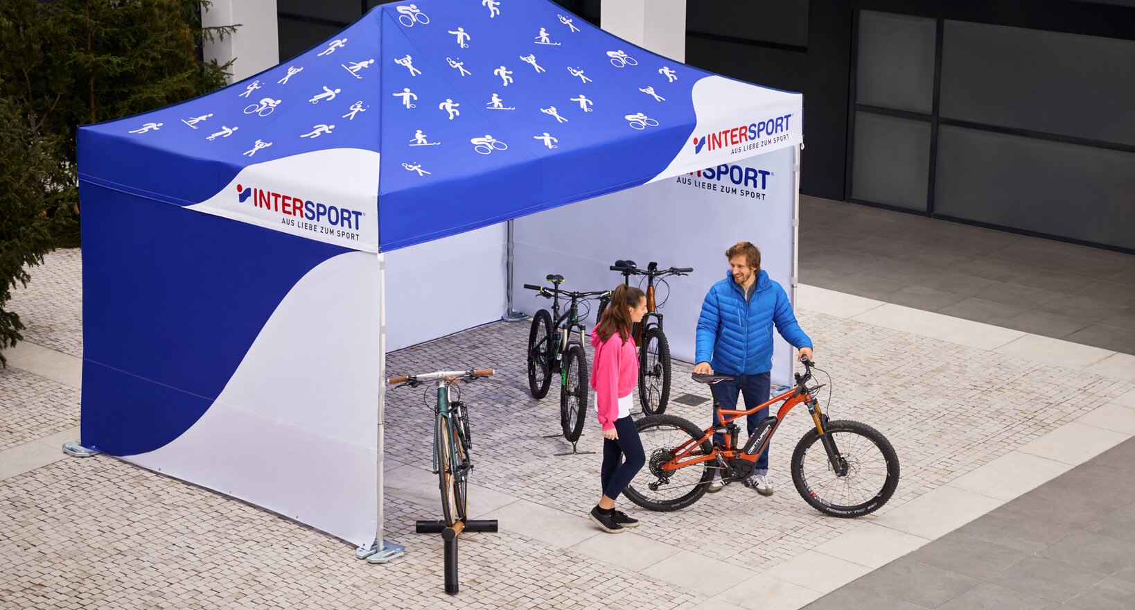 The promotion tent from Intersport is a 4.5x3 m folding gazebo. It is printed all over in white and blue. Various sport icons can be seen on the roof. The salesman shows the customer the bicycle.