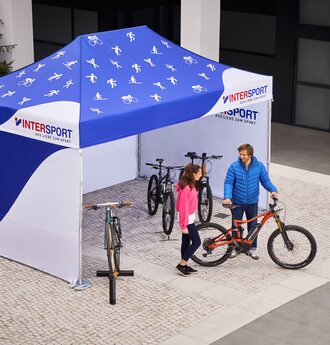 Il gazebo promozionale di Intersport è un gazebo pieghevole di 4,5x3 m. È stampato interamente in bianco e blu. Sul tetto sono visibili varie icone sportive. Il venditore mostra al cliente la bicicletta.