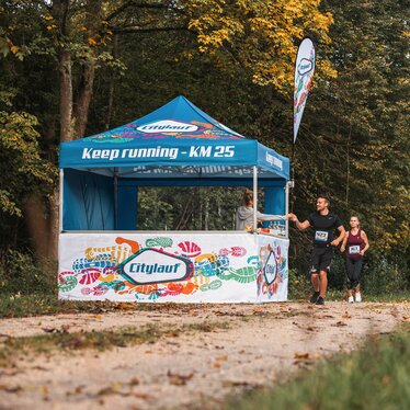 Gazebo per eventi blu 3x3m con stampa personalizzata Citylauf, con bandiera personalizzata, parete laterale chiusa e parete laterale a mezza altezza con bancone utilizzato come punto di ristoro a una corsa cittadina