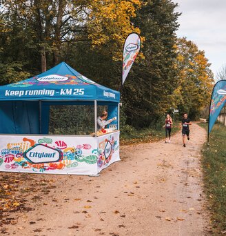 Das blau-weiße Eventzelt dient als Versorgungsstation bei einem Marathon. 2 Läufer laufen gerade am Eventzelt vorbei.