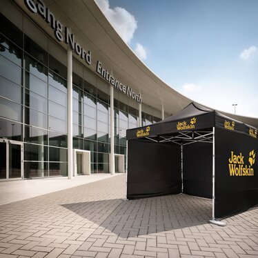 Das schwarze Messe- und Promotionzelt von Jack Wofskin ist ein 3x3 m Faltpavillon mit 3 geschlossenen Seitenwänden. Das Logo ist gelb und der Faltpavillon steht vor dem Messegelände.
