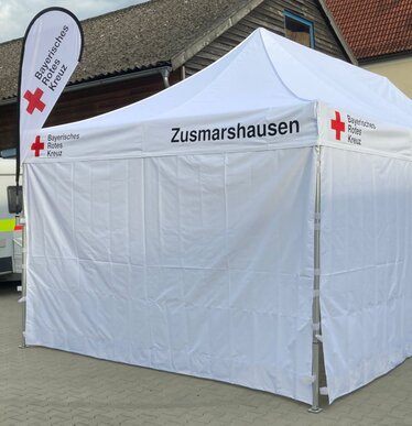Gazebo 8x4 bianco con pareti laterali chiuse e con finestra, personalizzato con scritta per Croce Rossa Bavarese e bandiera a goccia personalizzata con logo