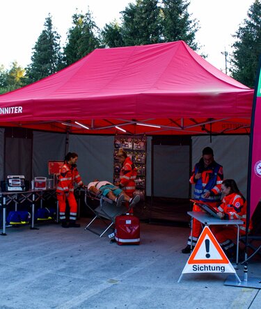 Rotes 6x4 Sanitätszelt mit grauen Rescue-Seitenwänden und zwei personalisierten Tropfenflaggen mit Logo, eingesetzt für eine Notfallsimulation auf einer Messe.