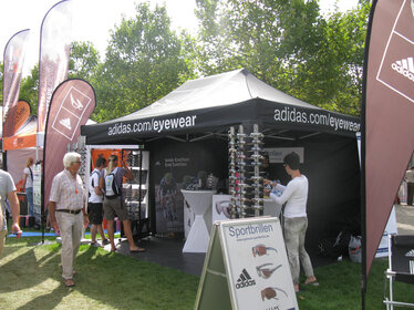 Gazebo pieghevole nero 3x4,5 personalizzato con stampa logo Adidas eywear con pareti laterali chiuse nere. Bandiere a goccia pubblicitarie bordeaux con logo. Fiera all'aperto con esposizione occhiali adidas. Ecotent