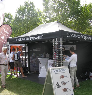 Gazebo pieghevole nero 3x4,5 personalizzato con stampa logo Adidas eywear con pareti laterali chiuse nere. Bandiere a goccia pubblicitarie bordeaux con logo. Fiera all'aperto con esposizione occhiali adidas. Ecotent