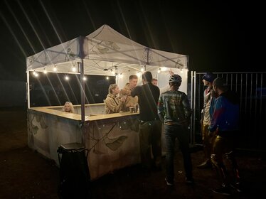 Gazebo pieghevole Ecotent® personalizzato con stampa foglie, utilizzato come bar all’aperto di notte con illuminazione e persone che si servono drink.