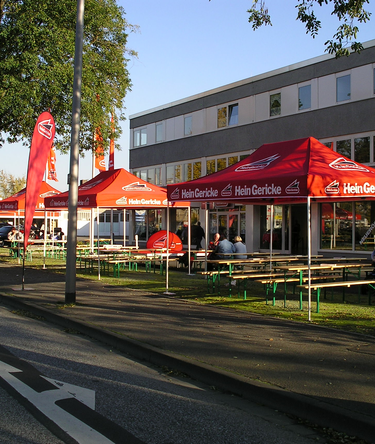 Gazebo pieghevoli promozionali rossi 4,5x3 personalizzati con logo Hein Gericke per evento all'esterno di un negozio. Ci sono felle bandiere pubbliciatrie a goccia personalizzate. Ecotent