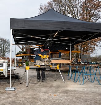 Gazebo nero 4x4m da lavoro con strisce led ripara un falegname che lavora in un cantiere edilizo vicino a un furgone